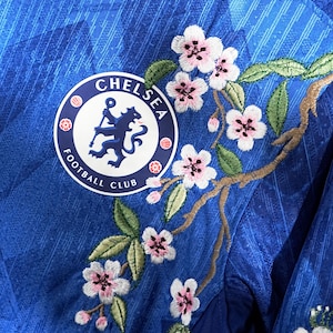 May include: A close-up of a blue Chelsea Football Club jersey with floral embroidery. The jersey displays the club's logo and a design of pink cherry blossoms and green leaves.