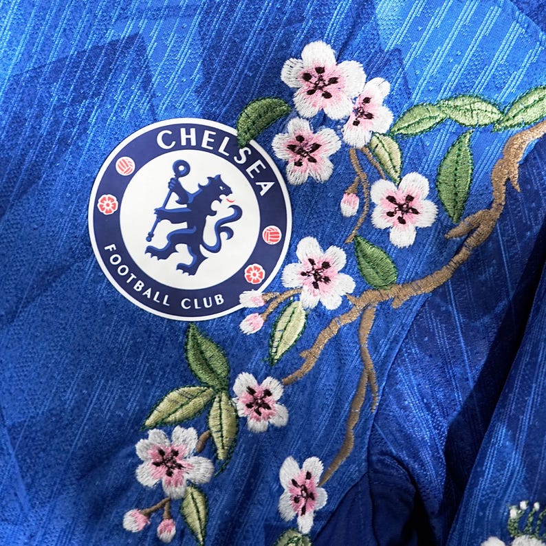 May include: A close-up of a blue Chelsea Football Club jersey with floral embroidery. The jersey displays the club's logo and a design of pink cherry blossoms and green leaves.