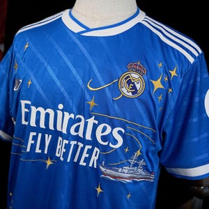 Puede incluir: Una camiseta de f&uacute;tbol azul real con detalles blancos, que presenta el escudo del equipo, estrellas doradas y el texto "Emirates FLY BETTER". La camiseta tiene cuello en V y rayas blancas en los hombros. Un barco est&aacute; bordado en la parte inferior.