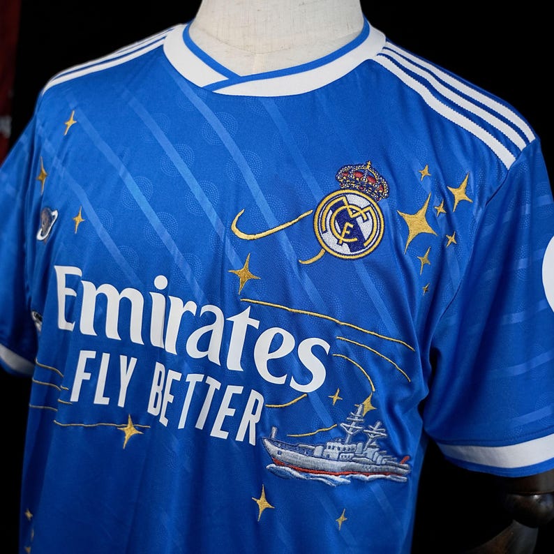Puede incluir: Una camiseta de f&uacute;tbol azul real con detalles blancos, que presenta el escudo del equipo, estrellas doradas y el texto "Emirates FLY BETTER". La camiseta tiene cuello en V y rayas blancas en los hombros. Un barco est&aacute; bordado en la parte inferior.