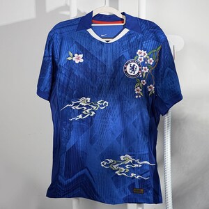 May include: A blue football shirt with a V-neck and short sleeves. The shirt has a team logo, floral embroidery, and cloud-like designs. The shirt is hanging on a white hanger.