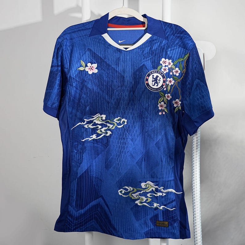 May include: A blue football shirt with a V-neck and short sleeves. The shirt has a team logo, floral embroidery, and cloud-like designs. The shirt is hanging on a white hanger.