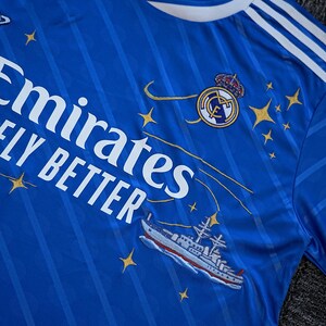 Puede incluir: Camiseta de f&uacute;tbol azul real con rayas blancas en los hombros y el texto "Emirates FLY BETTER". Presenta un escudo del equipo, detalles de estrellas y un dibujo de un barco.