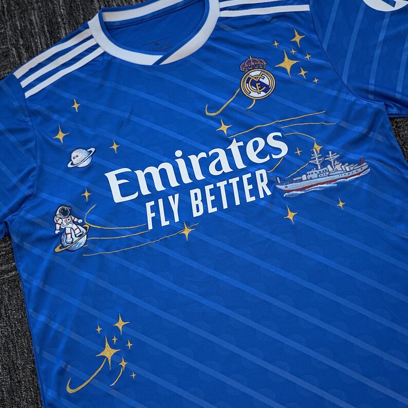 Puede incluir: Una camiseta de f&uacute;tbol azul con rayas blancas en los hombros y cuello blanco. La camiseta muestra el texto "Emirates FLY BETTER" y un logotipo del equipo. Tiene ilustraciones de una nave espacial, un astronauta y estrellas.