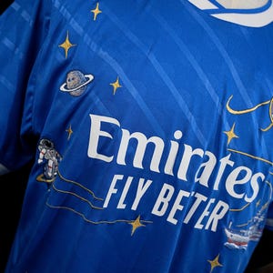 Puede incluir: Una camiseta azul real con el texto "Emirates FLY BETTER" en blanco. La camiseta presenta estrellas doradas bordadas, un planeta, un astronauta y un dise&ntilde;o de barco. El cuello es blanco y azul.