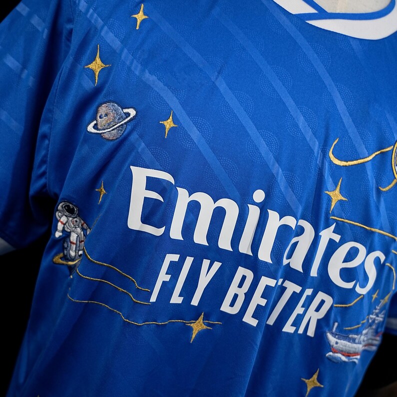Puede incluir: Una camiseta azul real con el texto "Emirates FLY BETTER" en blanco. La camiseta presenta estrellas doradas bordadas, un planeta, un astronauta y un dise&ntilde;o de barco. El cuello es blanco y azul.