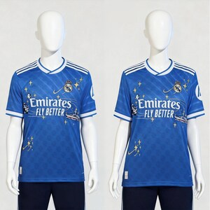 Puede incluir: Dos camisetas de f&uacute;tbol azules con detalles blancos y el texto "Emirates FLY BETTER." Las camisetas presentan un escudo, estrellas y una ilustraci&oacute;n de barco. Las camisetas se muestran en maniqu&iacute;es.