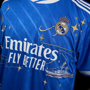Puede incluir: Camiseta de f&uacute;tbol azul real con detalles blancos y bordados dorados. La camiseta muestra el texto "Emirates FLY BETTER" y un escudo del equipo con una corona. Tambi&eacute;n hay gr&aacute;ficos de estrellas y barcos dorados. La camiseta tiene rayas blancas en los hombros.