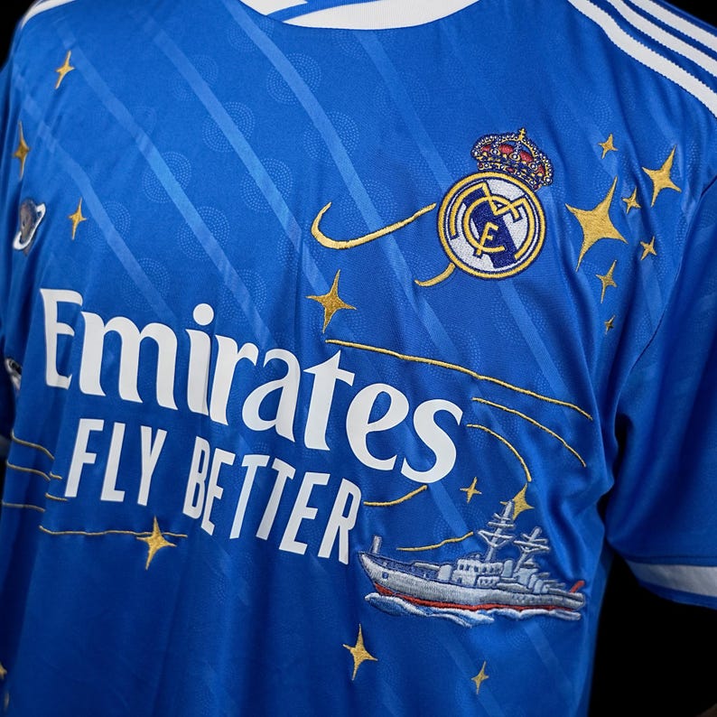 Puede incluir: Camiseta de f&uacute;tbol azul real con detalles blancos y bordados dorados. La camiseta muestra el texto "Emirates FLY BETTER" y un escudo del equipo con una corona. Tambi&eacute;n hay gr&aacute;ficos de estrellas y barcos dorados. La camiseta tiene rayas blancas en los hombros.