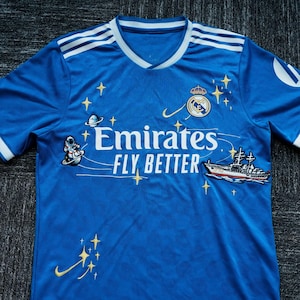 Puede incluir: Una camiseta de f&uacute;tbol azul con detalles blancos, que muestra el texto "Emirates FLY BETTER". La camiseta tiene cuello en V y el escudo del equipo. Presenta un astronauta, un barco y estrellas como elementos decorativos.