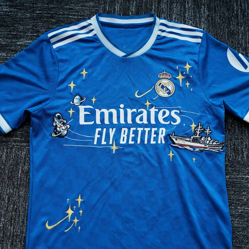Puede incluir: Una camiseta de f&uacute;tbol azul con detalles blancos, que muestra el texto "Emirates FLY BETTER". La camiseta tiene cuello en V y el escudo del equipo. Presenta un astronauta, un barco y estrellas como elementos decorativos.