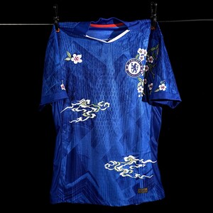 May include: A royal blue football shirt featuring white floral and cloud patterns. The shirt has a V-neck and the Chelsea Football Club emblem. It is suspended on a washing line against a black backdrop.
