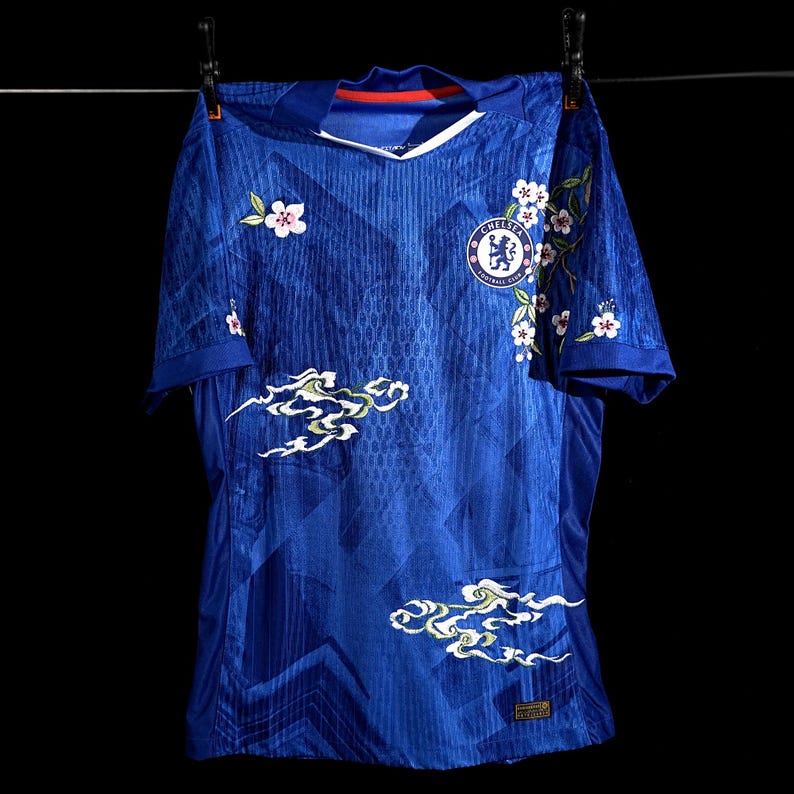 May include: A royal blue football shirt featuring white floral and cloud patterns. The shirt has a V-neck and the Chelsea Football Club emblem. It is suspended on a washing line against a black backdrop.