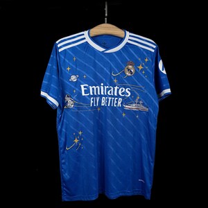 Puede incluir: Camiseta de f&uacute;tbol azul con rayas blancas en los hombros y las mangas. La camiseta presenta el texto "Emirates FLY BETTER" y un escudo del equipo. Tiene un dise&ntilde;o de estrellas, planetas y una nave espacial.