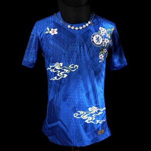 May include: A blue Chelsea FC football shirt with white floral and cloud embroidery. The shirt features the team's logo on the chest and a decorative necklace. The shirt is displayed on a black background.