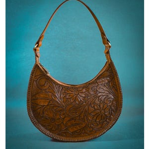 May include: A brown leather handbag with a floral embossed design. The crescent-shaped bag features a long, thin strap and is secured with metal hardware. The bag's surface is intricately carved with flowers and leaves.