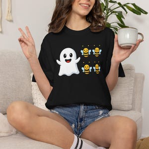 May include: A black short-sleeve t-shirt featuring a graphic print of a white cartoon ghost with a smiling face positioned next to four small yellow and black striped bees with light blue wings. The shirt is paired with blue denim shorts.