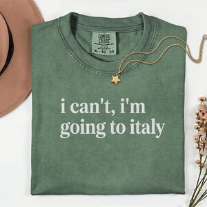 May include: Green Comfort Colors t-shirt with the text "i can't, i'm going to italy" in white. A gold necklace with a star pendant rests on the shirt. A brown hat and flowers are also in the image.