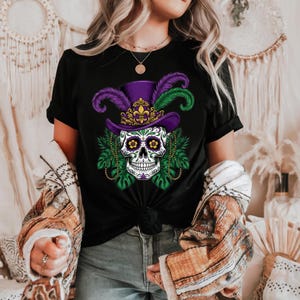 May include: Black t-shirt featuring a sugar skull design with a purple top hat adorned with feathers and a gold fleur-de-lis. The skull has floral eyes and is surrounded by green leaves and beaded necklaces. A festive design.