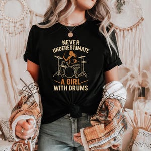 Puede incluir: Una camiseta negra de manga corta con un estampado gráfico en color tostado. El diseño muestra la silueta de una persona tocando la batería con el texto "NEVER UNDERESTIMATE A GIRL WITH DRUMS" en una fuente sans-serif negrita y mayúscula.
