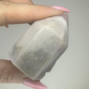 May include: A faceted crystal point with a light purple tip and a grey and white body. The crystal is held by a person's hand, showcasing its natural texture and colour variations. The crystal's shape is geometric, with visible facets.