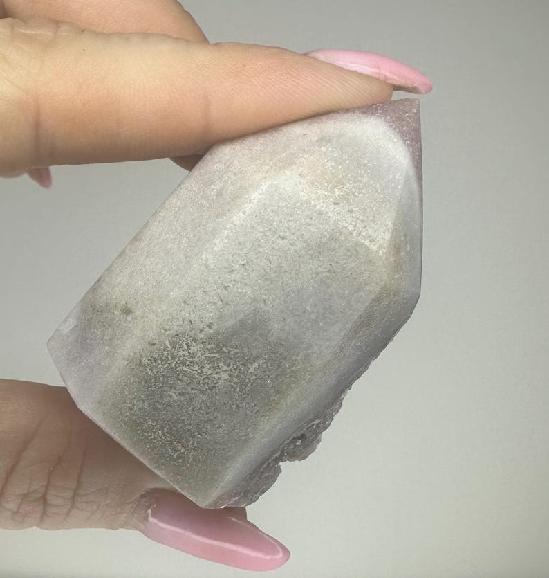 May include: A faceted crystal point with a light purple tip and a grey and white body. The crystal is held by a person's hand, showcasing its natural texture and colour variations. The crystal's shape is geometric, with visible facets.