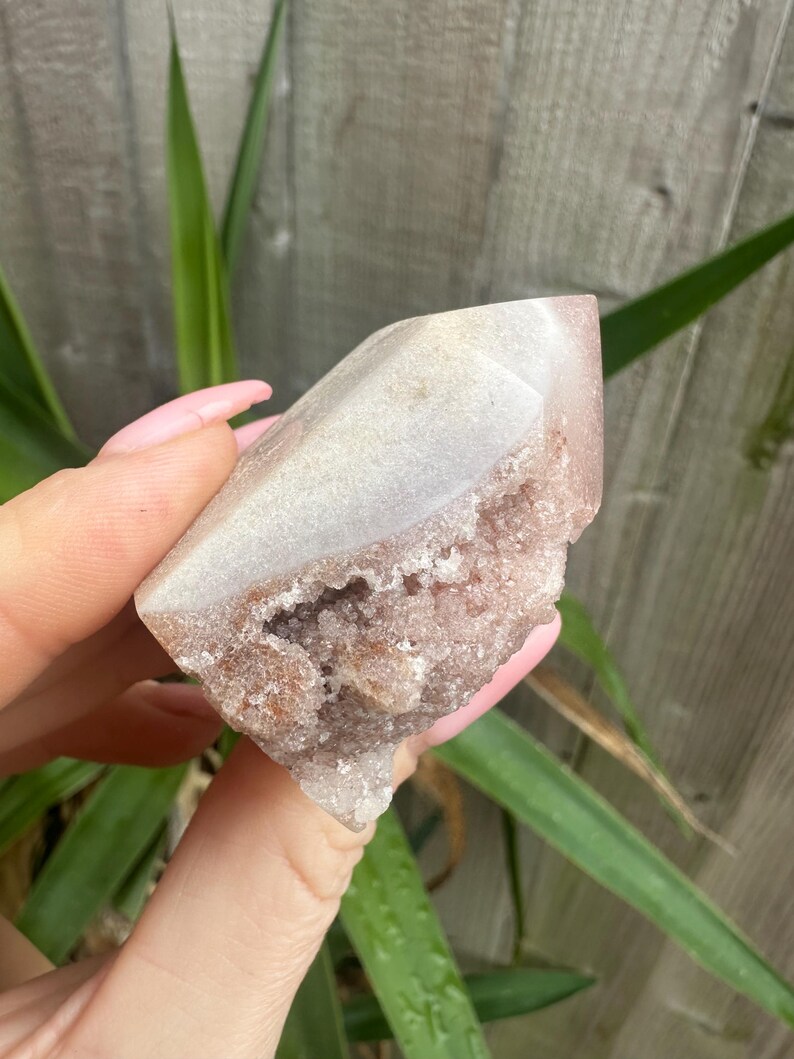 May include: A light pink and white crystal cluster with a pointed top. The crystal has a rough, textured surface with a cavity in the centre. The crystal is held in a hand, with green leaves and a wooden fence in the background.