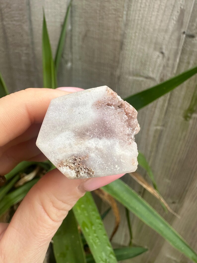May include: A hexagonal crystal in shades of white and pale purple. The crystal has a rough, textured edge with a brown tone. The crystal is held in a hand, with green leaves visible in the background.