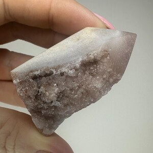 May include: A natural crystal formation, predominantly light purple with a white, crystalline top. The rough, textured surface reveals a complex internal structure. The crystal is held in a hand, highlighting its unique shape and colour.