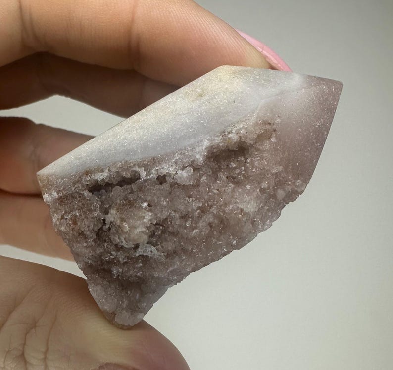 May include: A natural crystal formation, predominantly light purple with a white, crystalline top. The rough, textured surface reveals a complex internal structure. The crystal is held in a hand, highlighting its unique shape and colour.