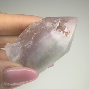 May include: A light purple crystal point with a rough, textured edge. The crystal has a smooth, polished surface that transitions from a pale lavender to a clear, translucent tip. The crystal is held in a hand.