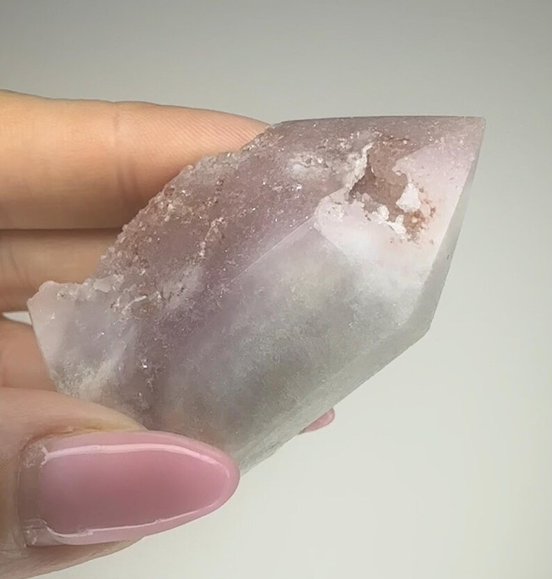 May include: A light purple crystal point with a rough, textured edge. The crystal has a smooth, polished surface that transitions from a pale lavender to a clear, translucent tip. The crystal is held in a hand.