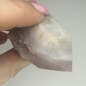 May include: A crystal point with a colour gradient, shifting from a pale, translucent white to a soft lavender. The base has a rough texture, while the top is smooth and faceted. The crystal is held by a hand.