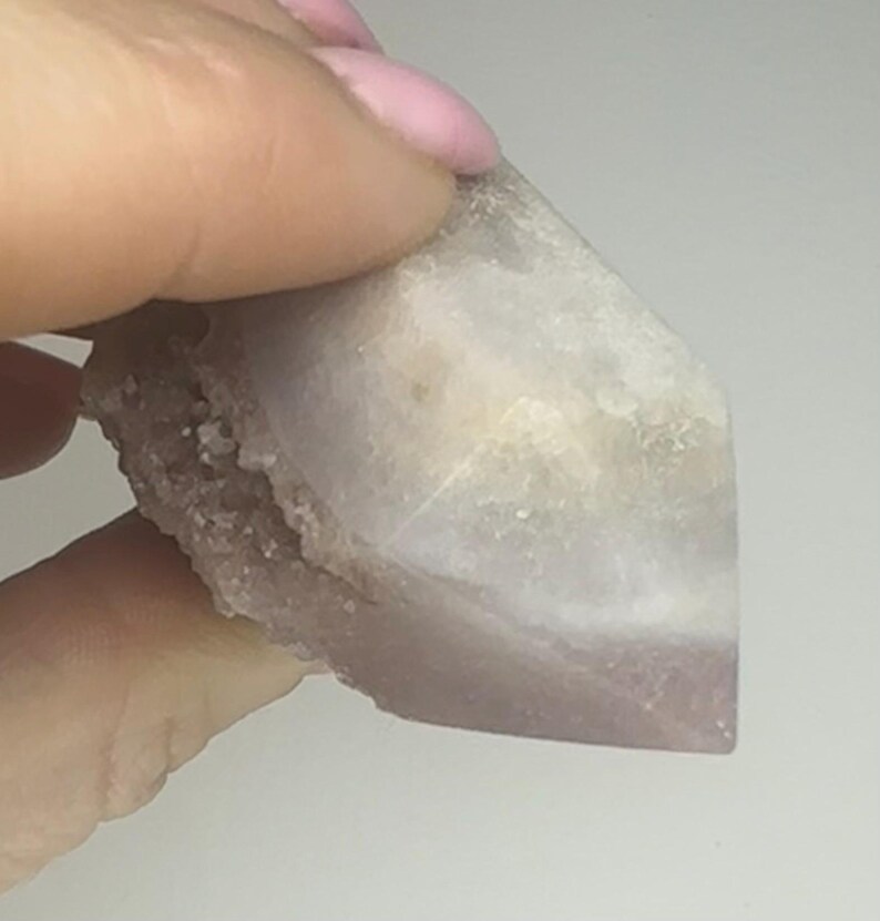 May include: A crystal point with a colour gradient, shifting from a pale, translucent white to a soft lavender. The base has a rough texture, while the top is smooth and faceted. The crystal is held by a hand.