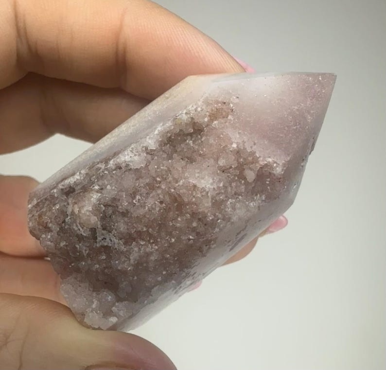 May include: A light pink crystal point with a rough, textured base and a smooth, polished tip. The crystal has a translucent quality, with areas of darker, reddish-brown inclusions. The crystal is held in a hand.