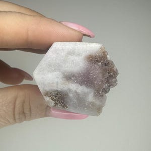 May include: A hexagonal crystal in shades of white, lavender, and brown. The crystal is held between fingers, highlighting its natural, rough texture and colour variations. The edges are uneven, adding to its organic appeal. The crystal is a natural stone.