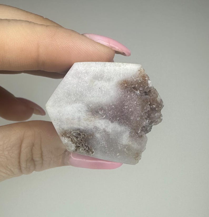 May include: A hexagonal crystal in shades of white, lavender, and brown. The crystal is held between fingers, highlighting its natural, rough texture and colour variations. The edges are uneven, adding to its organic appeal. The crystal is a natural stone.