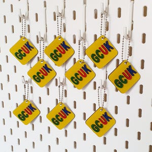 Peut inclure: Plusieurs porte-clés jaunes en forme de losange avec les lettres "GCUK" en vert, bleu et rouge. Les porte-clés sont suspendus à des chaînes à billes argentées et des crochets blancs sur un fond de panneau perforé blanc.