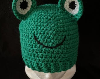 May include: A green crocheted frog hat with white and black eye details and a smiling mouth. The hat is designed to fit a head and is made of a soft, textured yarn. The frog hat is a fun and whimsical accessory.