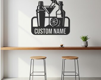 May include: Black metal wall art featuring cocktail-themed designs, including a shaker, martini glass, bottle, and a lemon slice. The text "CUSTOM NAME" is displayed on a rectangular base. The art is mounted on a white wall above a wooden counter.