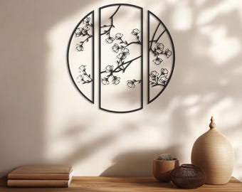 May include: Black metal wall art featuring a circular design with three panels. The art depicts a floral branch with delicate blossoms. The piece is mounted on a light-colored wall, with a wooden table in the foreground.