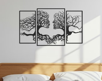Op de afbeelding: Een driedelig wandkunstwerk met een zwart metalen ontwerp van twee gezichten verweven met boomtakken. Het kunstwerk is gemonteerd op een witte muur boven een houten hoofdbord en wit beddengoed.