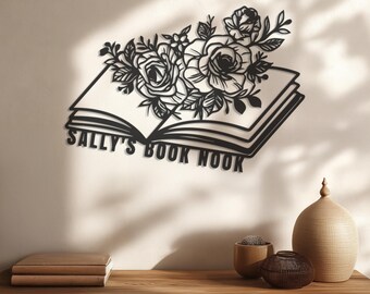 May include: Black metal wall art featuring an open book design with floral accents. The text "SALLY'S BOOK NOOK" is displayed across the open pages. The artwork is mounted on a light-colored wall, with decorative items on a wooden surface below.