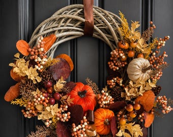Può includere: Una ghirlanda decorativa con una base di rami intrecciati decorata con zucche arancioni e bianche, pigne, foglie rosso scuro e dorate e grappoli di piccole bacche. La composizione è arricchita da nastri marroni e color crema che pendono.