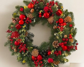 May include: A festive Christmas wreath with evergreen branches, red berries, and ornaments, including pinecones and round red baubles. The wreath is hanging on a white wall above a black table with a lamp and decorative items.
