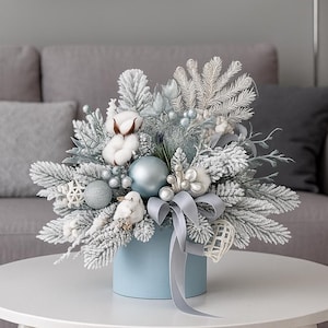 Könnte beinhalten: Festlicher Weihnachts-Tischschmuck in einem hellblauen quadratischen Behälter. Das Arrangement besteht aus gefrorenen Tannenzweigen, silbernen und blauen Ornamenten, Baumwollkugeln und einem grauen Band. Der Tischschmuck steht auf einem weißen Tisch.