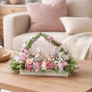 May include: A floral arrangement in a white rectangular container, featuring pink, white, and green flowers, with a small white bunny figurine. The arrangement is on a wooden coffee table in a living room setting.