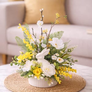 Könnte beinhalten: Ein Arrangement mit einem weißen Keramik-Hasen, gelben und weißen Blumen, grünem Laub und Zweigen. Die Anordnung befindet sich in einem weißen, gerippten Keramiktopf auf einer gewebten Matte. Der Tisch ist aus Marmor mit goldenen Beinen.