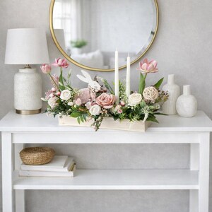 May include: An Easter-themed floral arrangement in a wooden box, with pink and white roses, greenery, and a bunny ornament. Two white candles and vases are on a white table, with a gold mirror in the background. The table is 100 cm long.