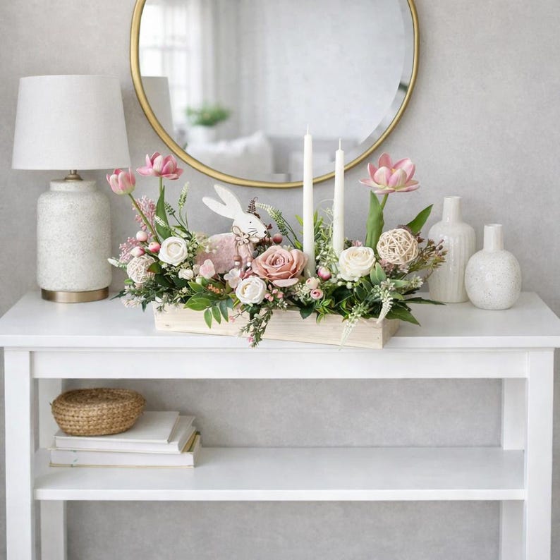 May include: An Easter-themed floral arrangement in a wooden box, with pink and white roses, greenery, and a bunny ornament. Two white candles and vases are on a white table, with a gold mirror in the background. The table is 100 cm long.